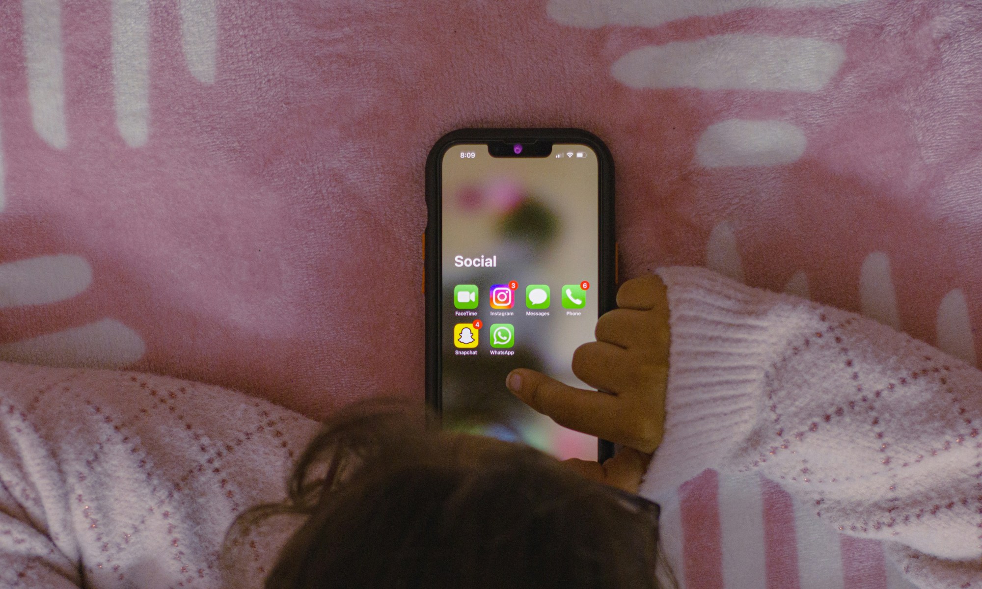 A human, most likely a kid, using a mobile phone pictured from above. Social apps shown on screen. Background is a pale pink blanket.