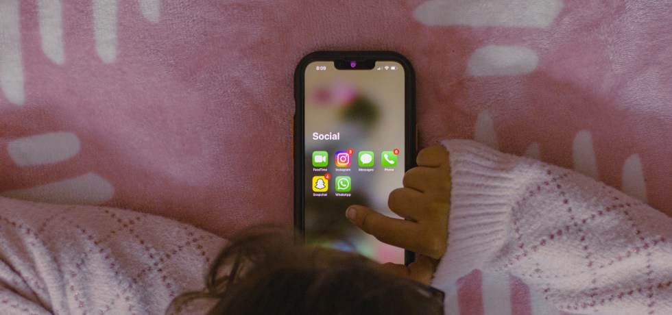 A human, most likely a kid, using a mobile phone pictured from above. Social apps shown on screen. Background is a pale pink blanket.